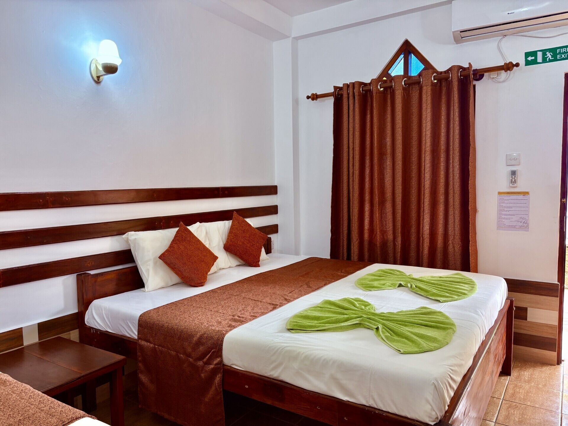 Habitación de hotel con cama de madera, mesita de noche, lámpara de pared, cortina y equipo de aire acondicionado.