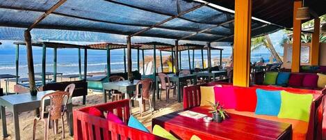 An outdoor seating area with tables and chairs, a wooden bench, and a view of the beach and ocean.