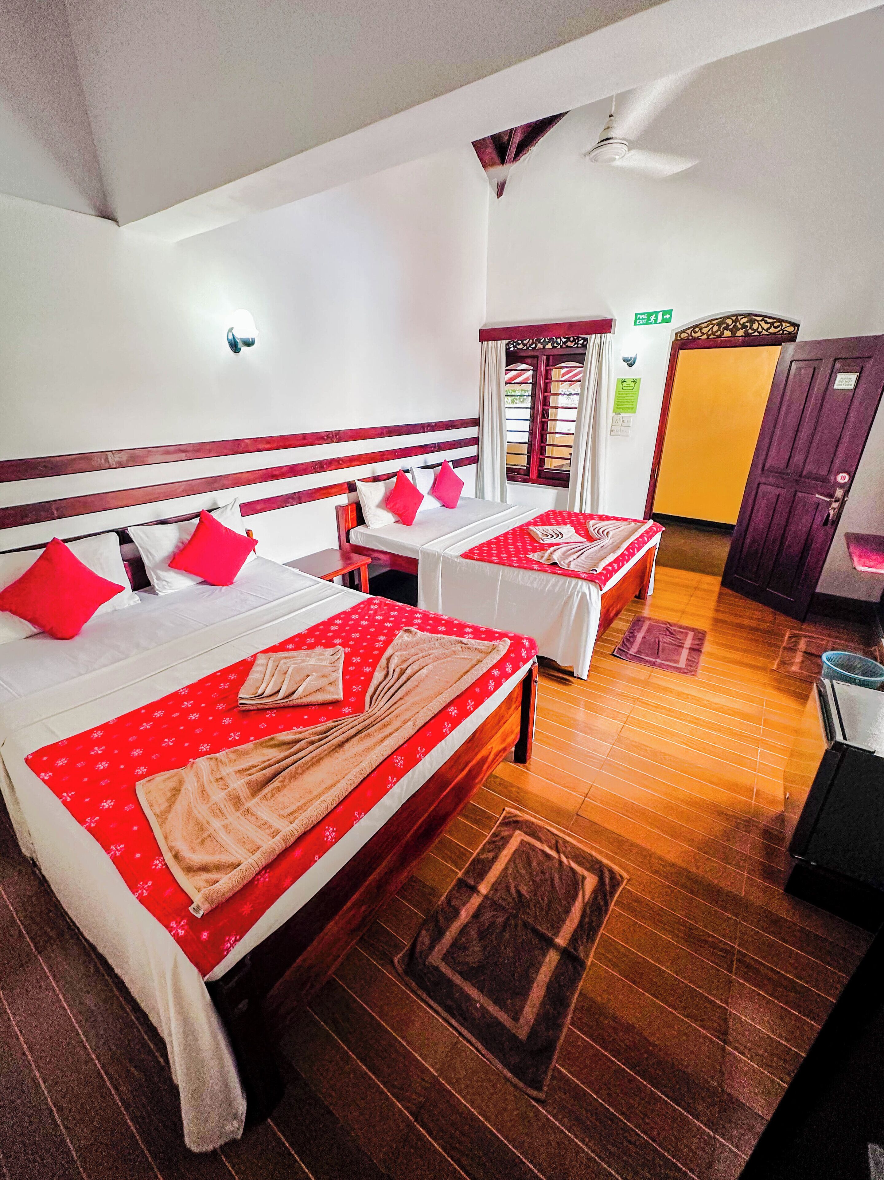 A hotel room with two beds, each with red and white bedding, wooden headboards, and a red floral bedspread. There is a wooden floor, a small table, and a door in the background.