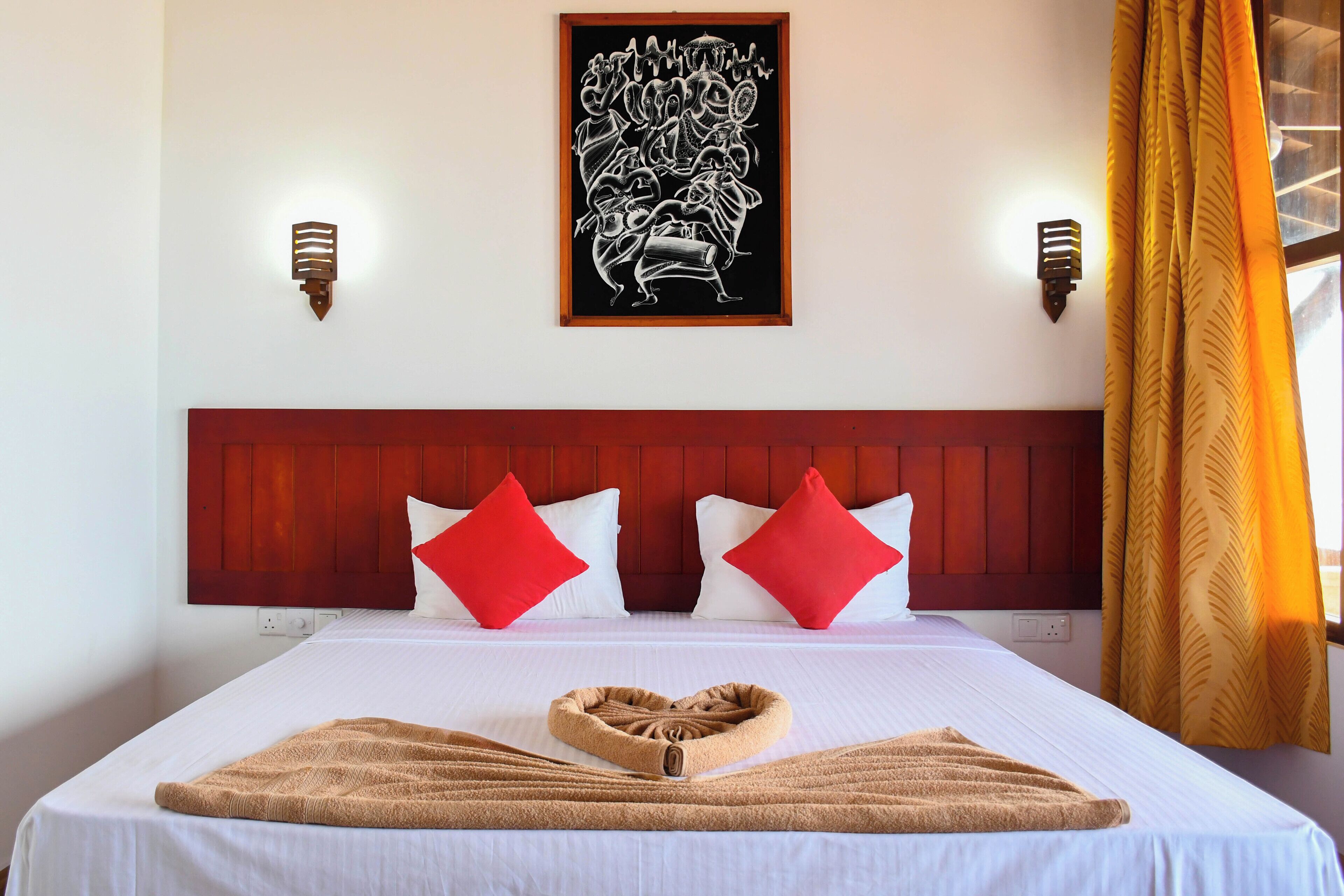 A hotel room with a large bed, red and white pillows, a wooden headboard, a framed artwork above the bed, and a window with yellow curtains.