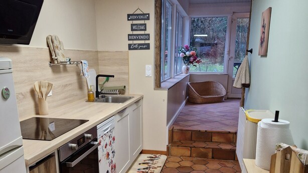 Ett kompakt kök med modern design, utrustat med en inbyggd ugn, en diskho och en mikrovågsugn. Köket har en bänkskiva i trä, en liten matplats med en bänk och en utsikt över trädgården genom stora fönster.