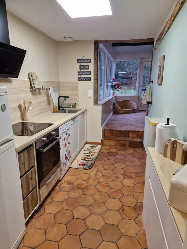 Una cocina compacta de diseño moderno, con horno empotrado, pileta y microondas. Cuenta con una encimera de madera, un pequeño comedor con banco y vistas al jardín a través de amplios ventanales.