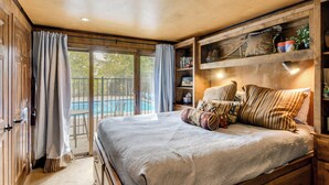 Un dormitorio con una cama grande, cabecera de madera, estante empotrado con objetos decorativos y vista a una piscina a través de una puerta corrediza de vidrio.
