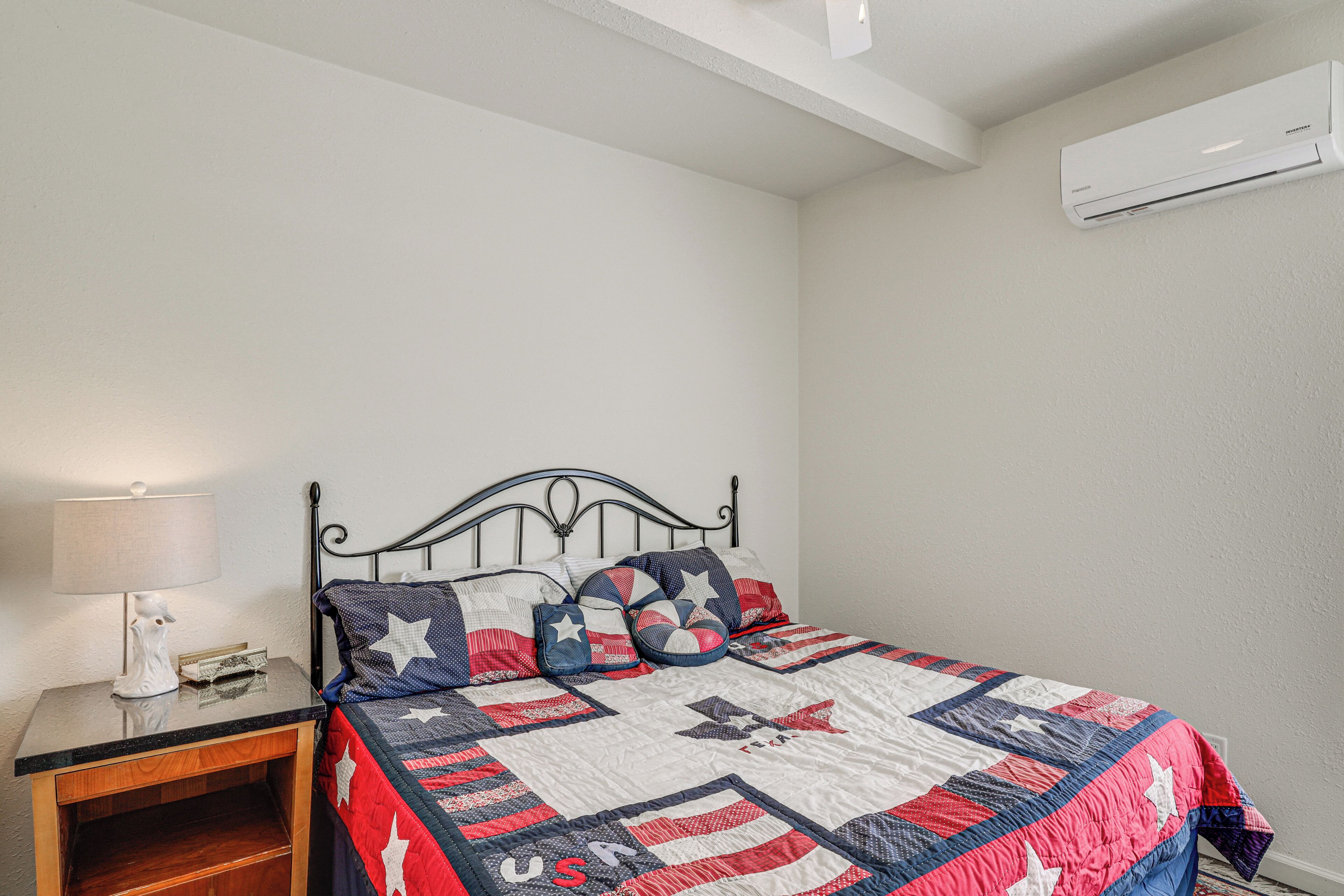 Un lit bien fait, recouvert d’une courtepointe à motifs étoilés, une table de chevet avec une lampe et un climatiseur fixé au mur.
