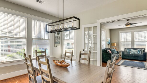 Un comedor con mesa y sillas de madera, una araña y ventanas amplias con persianas.
