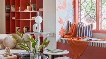 Un salón acogedor con una estantería roja, una mesa de centro redonda con libros y un jarrón, y un rincón con cojines junto a la ventana.