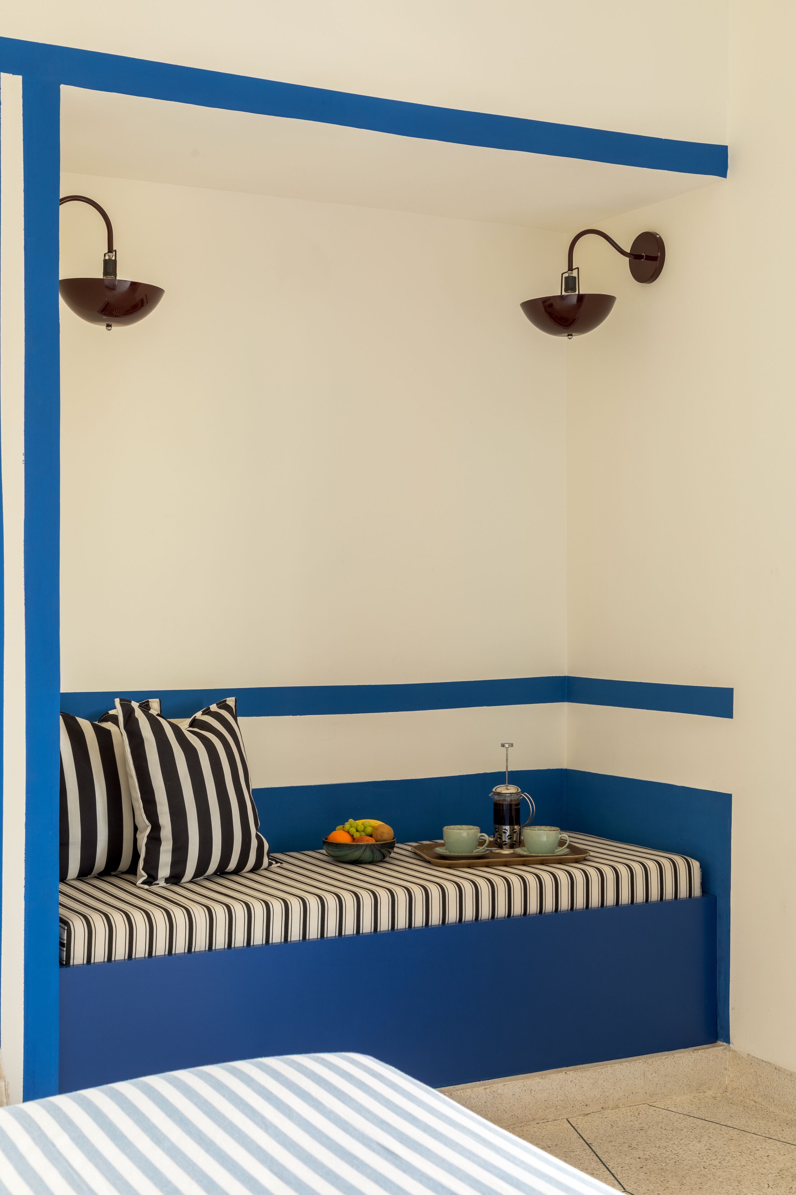 Um quarto com esquema de cores azul e branco, com um sofá listrado com almofadas, um tabuleiro com xícaras e frutas, e duas luminárias fixadas na parede.