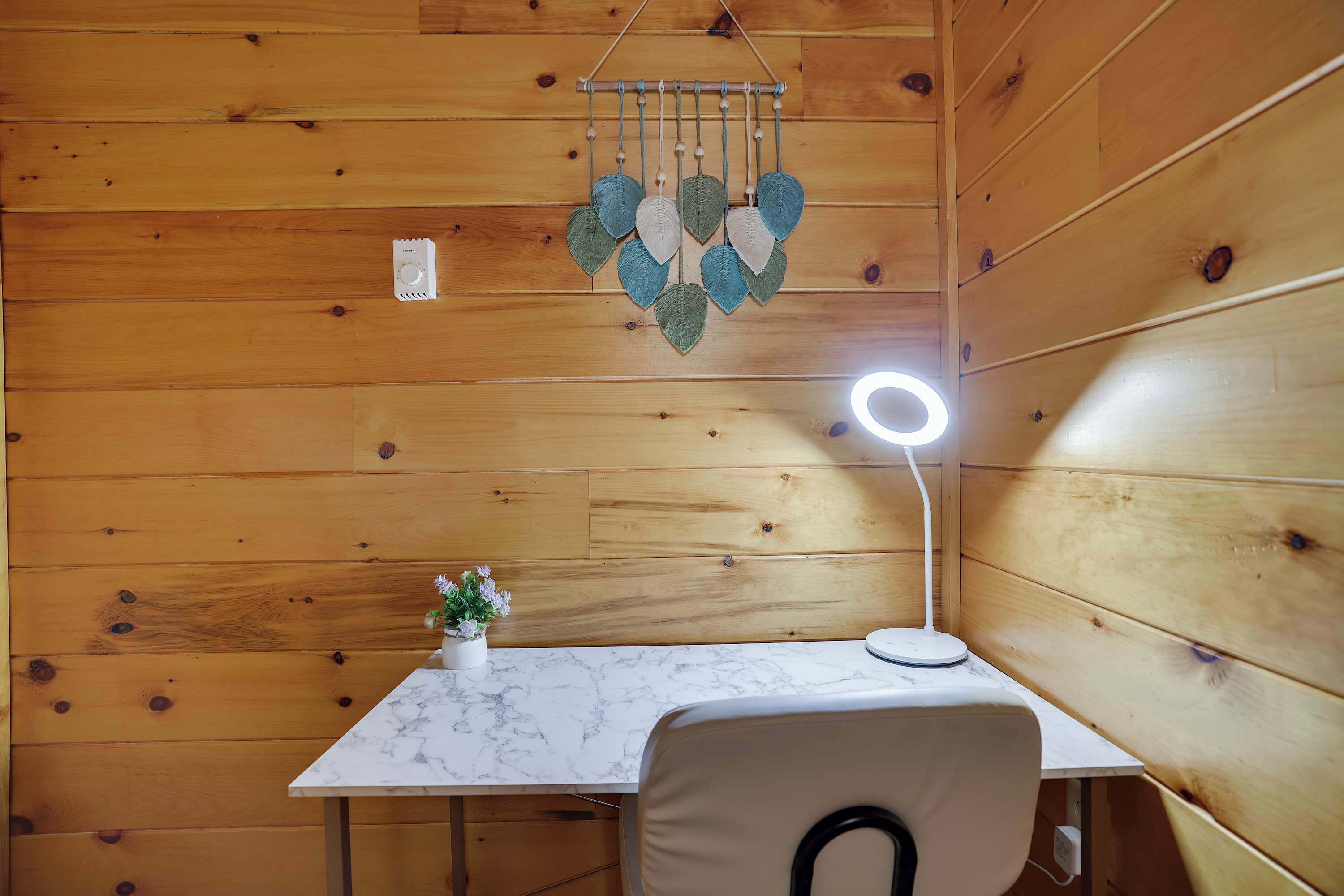 Interior de una cabaña de madera con una mesa de mármol, un adorno colgante de hojas, una pequeña planta en maceta y una lámpara de escritorio.