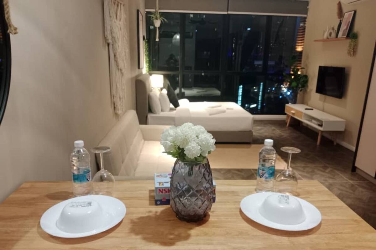 Un coin repas avec une table en bois, deux assiettes blanches, un vase avec des fleurs blanches et un canapé en arrière-plan.