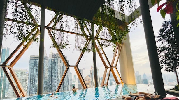 An indoor infinity pool with a cityscape view, surrounded by geometric metal structures and greenery.