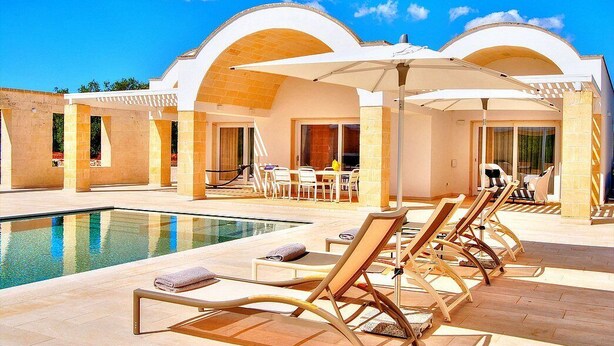 Ett område vid poolen med solstolar och parasoller, en pool och en byggnad med välvda fönster.