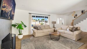 Un salon spacieux avec deux canapés beige, une table basse ronde et une grande plante en pot. On aperçoit un escalier au fond.