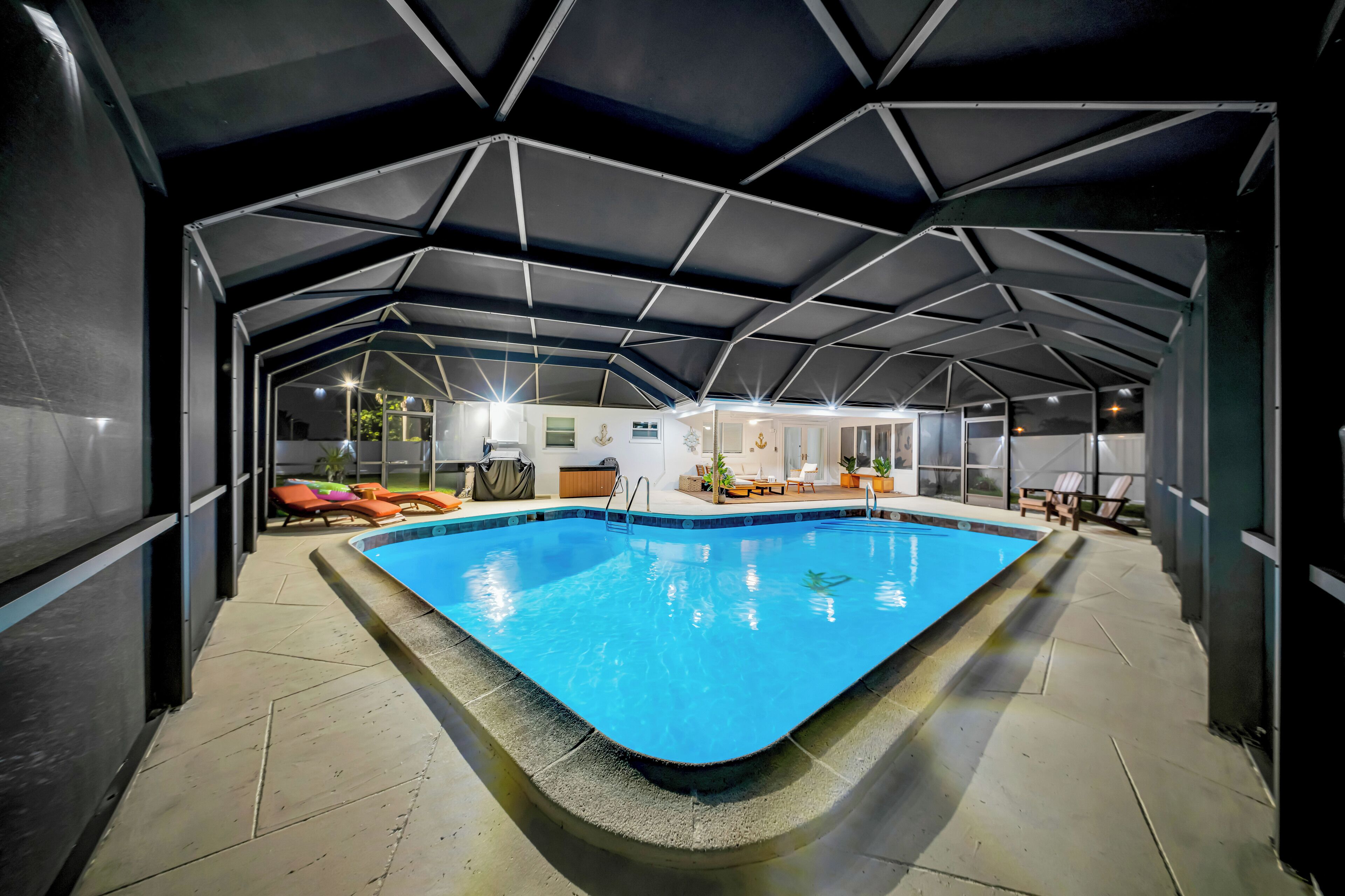 An indoor swimming pool with a geometric ceiling structure, surrounded by lounge chairs and a small seating area.