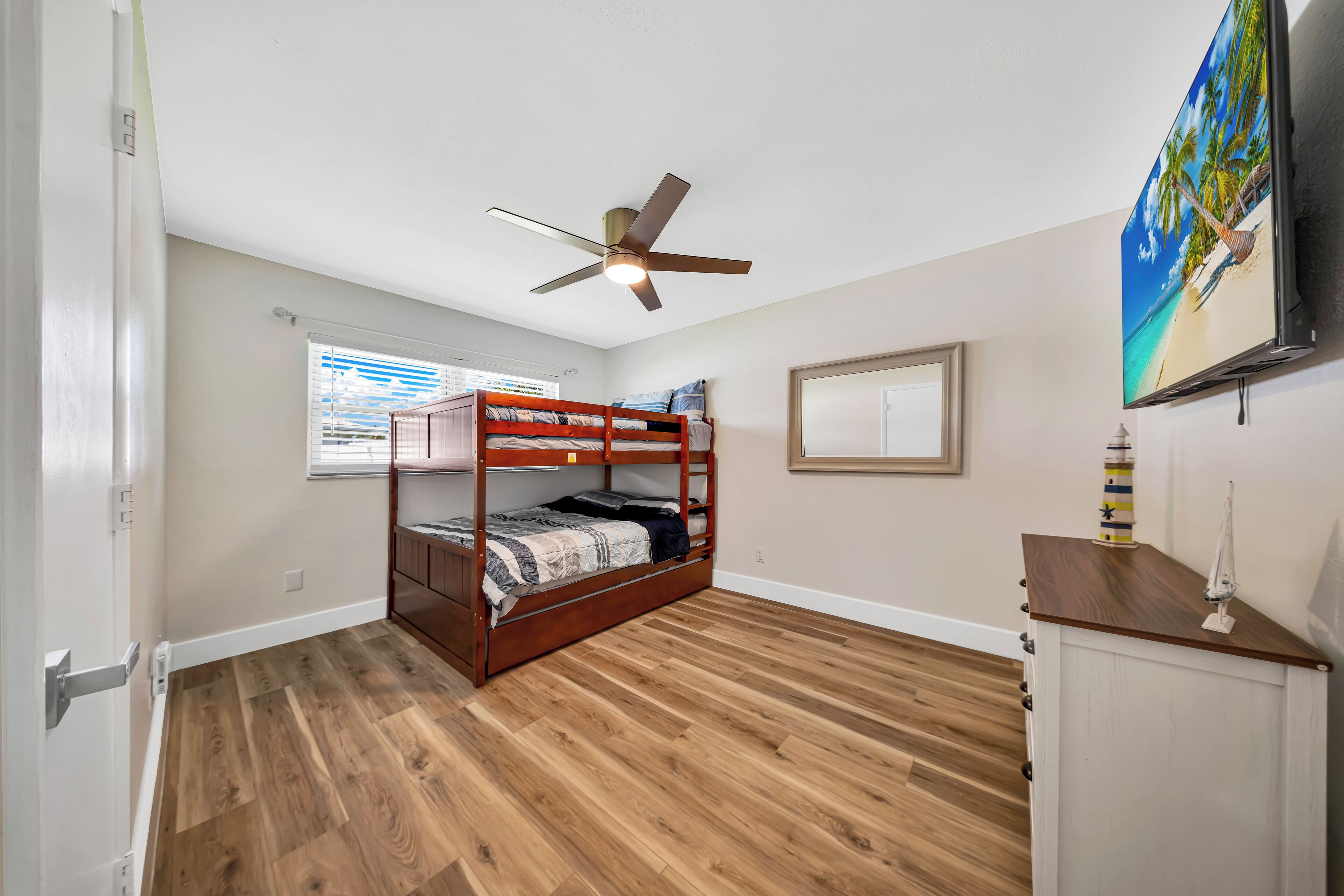 Une chambre avec un lit superposé, un ventilateur de plafond, un téléviseur fixé au mur et une fenêtre avec des stores.