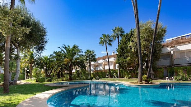 A swimming pool surrounded by palm trees and a building with a tiled roof. - Winahost Apartment Jávea (Jávea)