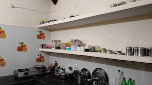 A kitchen with white tiled walls, a gas stove, and various kitchen utensils and containers on shelves and countertops. - Shree Shyam Paying Guest House (Varanasi)