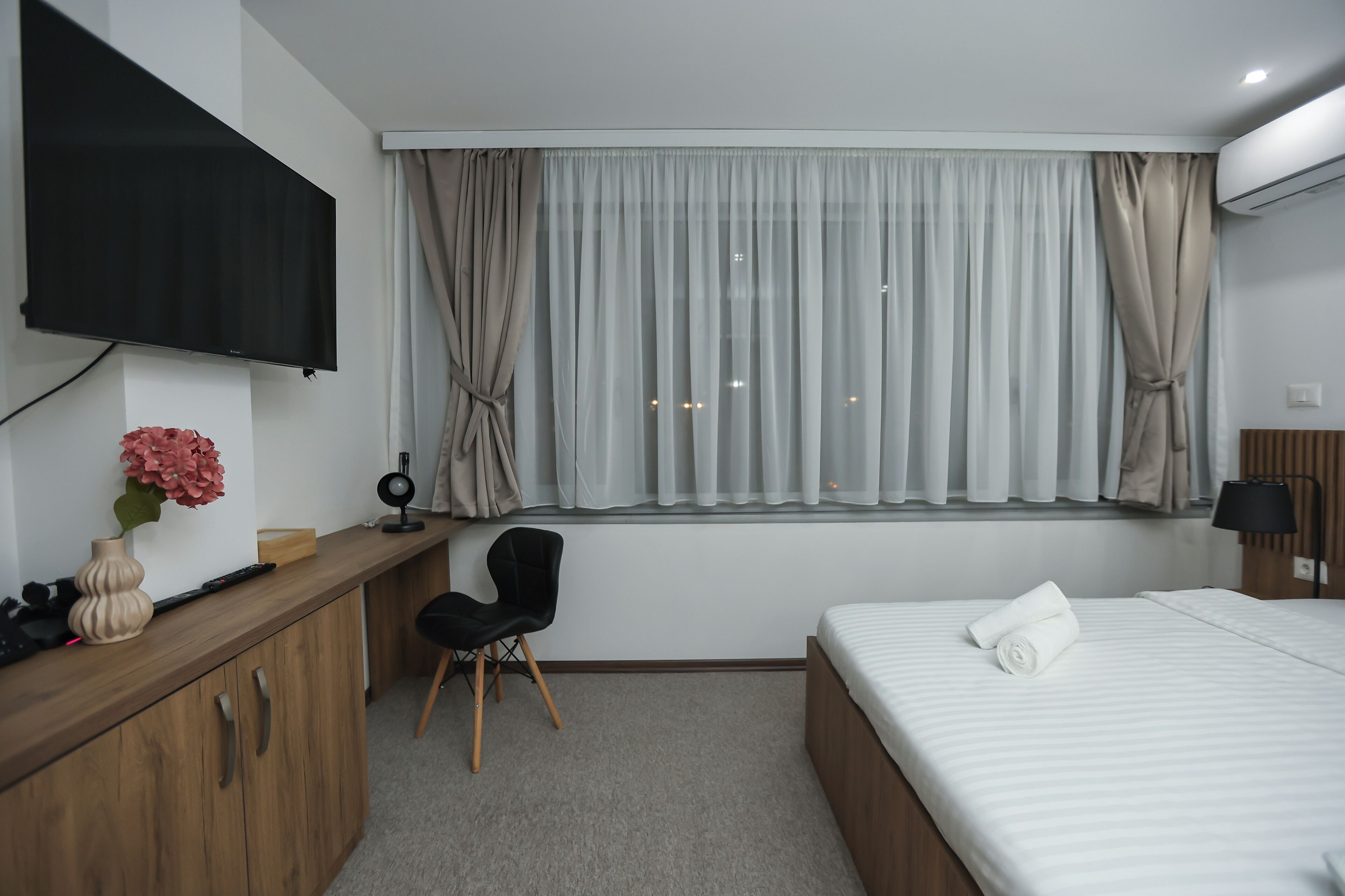 A hotel room with a bed, a desk with a chair, a TV mounted on the wall, and a window with curtains.