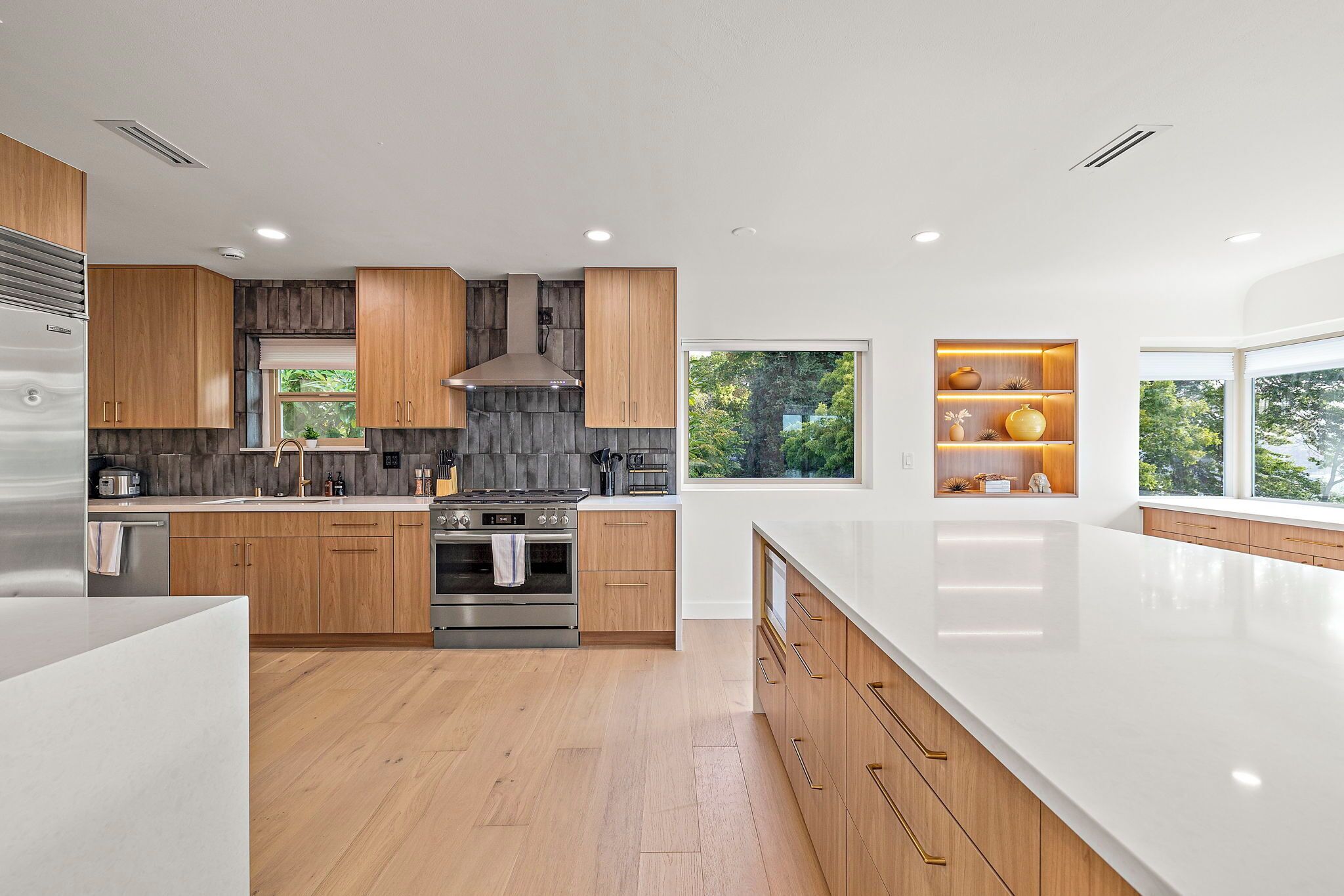 A modern kitchen with wooden cabinets, a stainless steel refrigerator, and a large window providing a view of greenery.