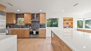 A modern kitchen with wooden cabinets, a stainless steel refrigerator, and a large window providing a view of greenery. - Sunlit & Spacious Seattle Home near Space Needle (Seattle)