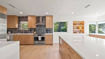 A modern kitchen with wooden cabinets, a stainless steel refrigerator, and a large window providing a view of greenery.