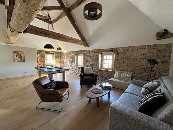 Un espacio tipo loft con vigas de madera a la vista, una mesa de billar, un sillón de cuero, un sofá y una pared de piedra.