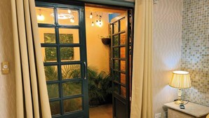 Una habitación de hotel con una cama, una mesita de noche con lámpara, una ventana con cortinas y una puerta que da a un balcón con plantas.