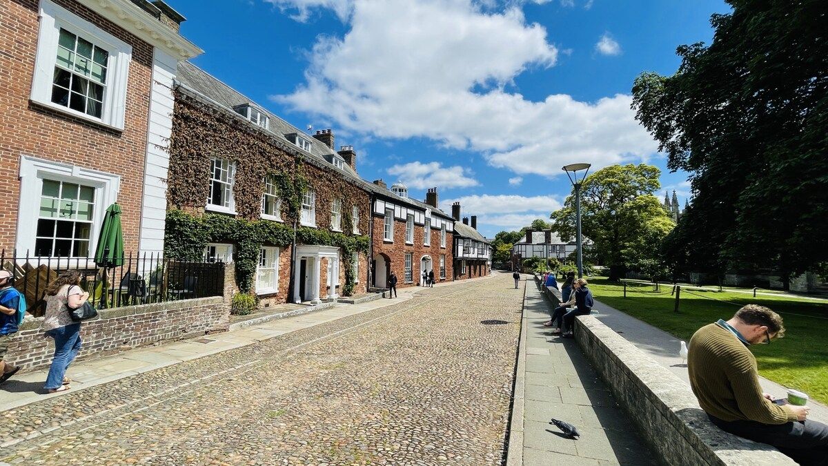 A cobblestone street lined with traditional brick buildings, a paved walkway, and a grassy area with benches.