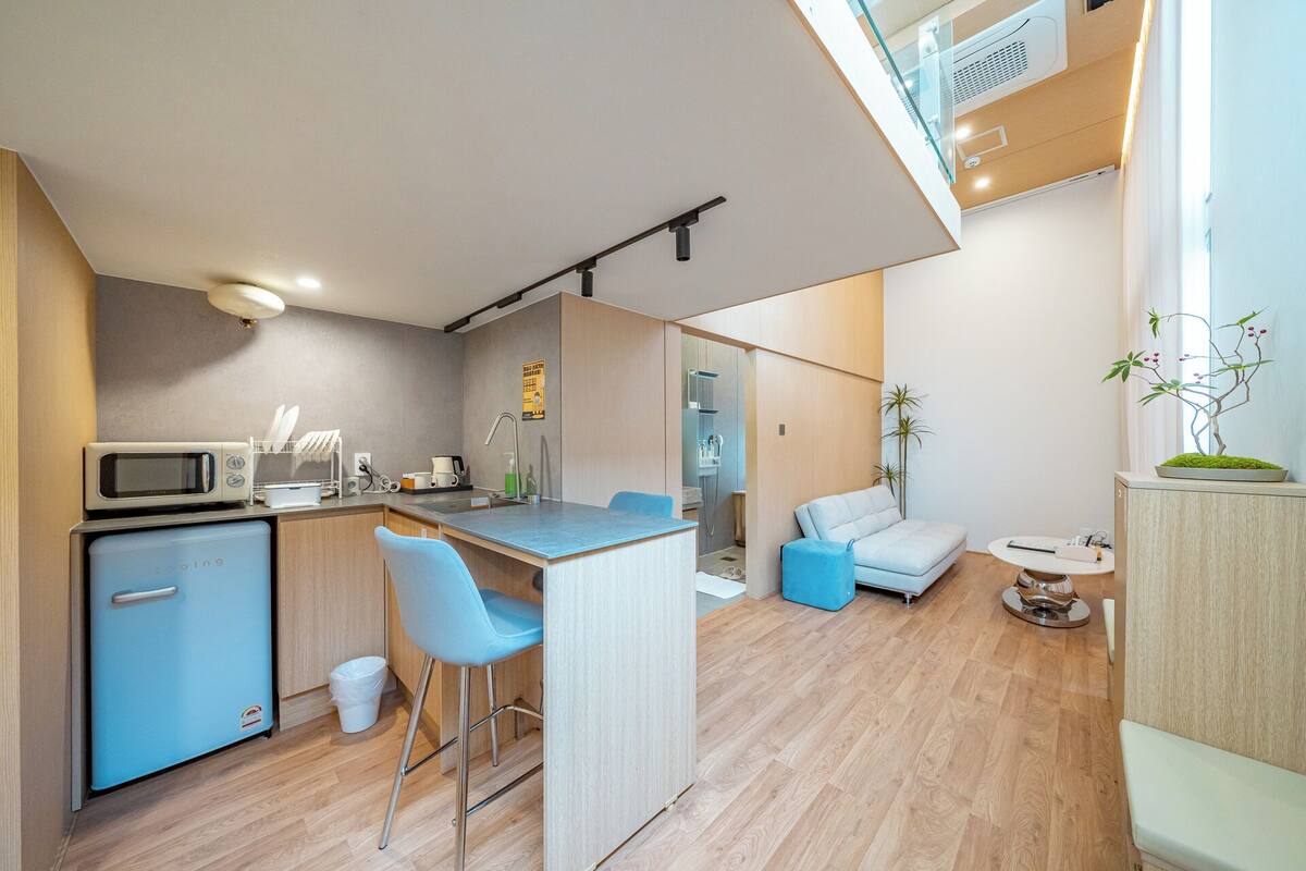 Cocina compacta con microondas, tostadora y pileta. Hay un refrigerador pequeño, un taburete y un sofá en la sala.