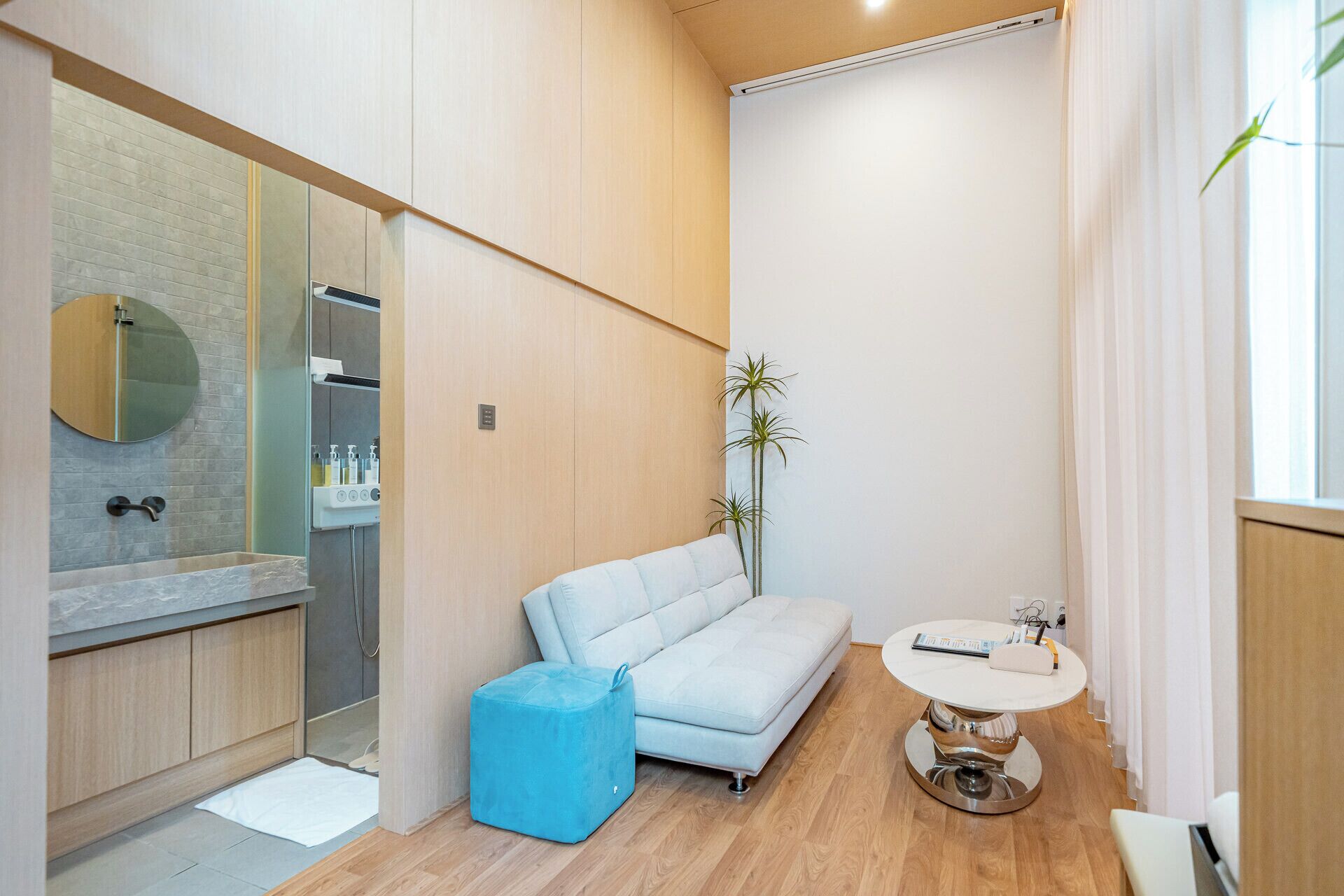A modern bathroom with a shower, a sink, and a mirror. There is a blue ottoman and a small round table with a book and a device.
