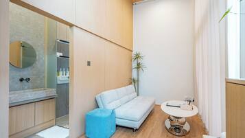 A modern bathroom with a shower, a sink, and a mirror. There is a blue ottoman and a small round table with a book and a device.