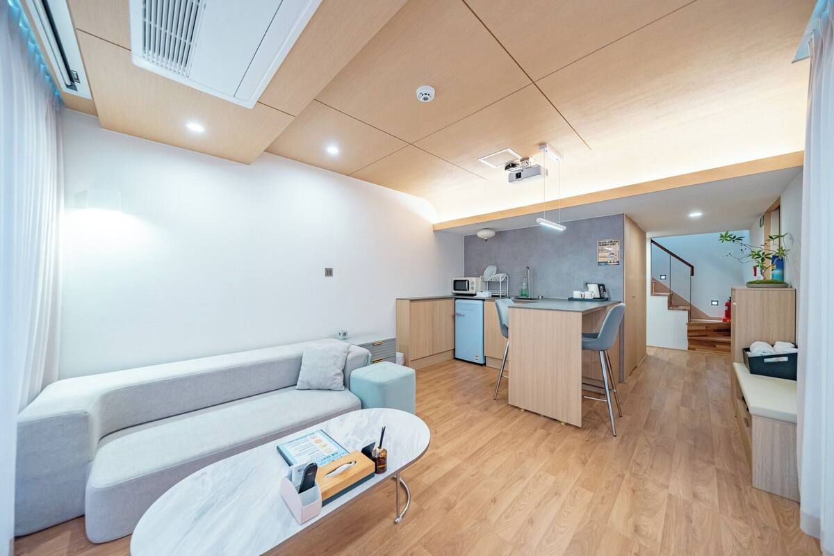 Un espacio habitable compacto con sofá, una pequeña mesa de comedor, kitchenette y una escalera que lleva a un nivel superior.