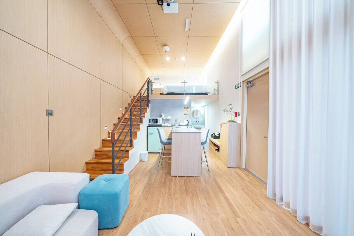 Una habitación de hotel moderna con una escalera de madera, una kitchenette, un comedor y una sala de estar con sofá y reposapiés.