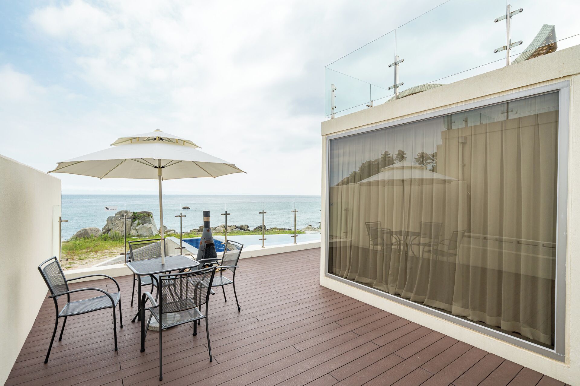 A balcony with a table and chairs, a large window with curtains, and a view of the sea.