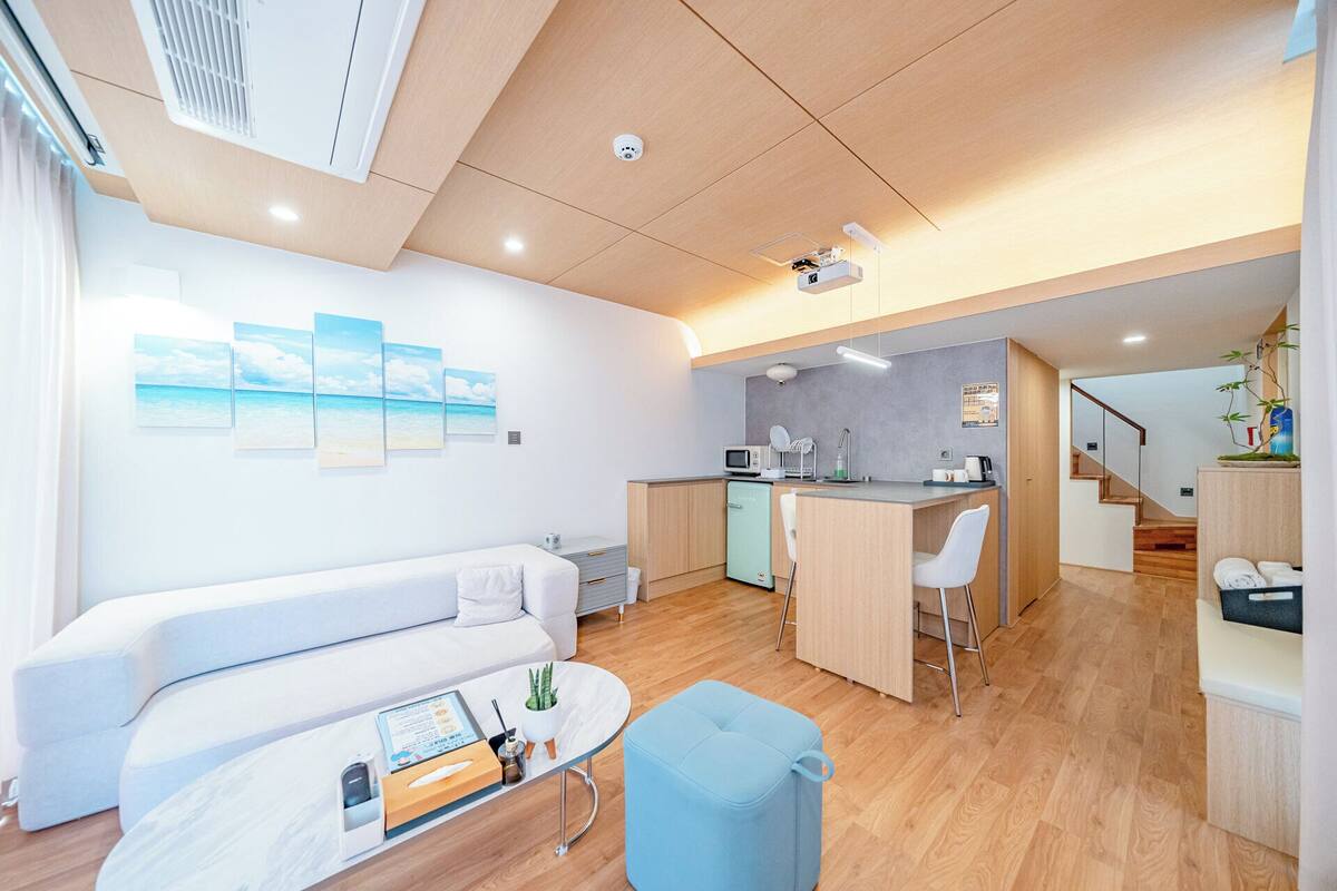 Una sala moderna con un sofá blanco, una mesita pequeña y un reposapiés azul. Hay una zona de cocina con un microondas y un lavabo.