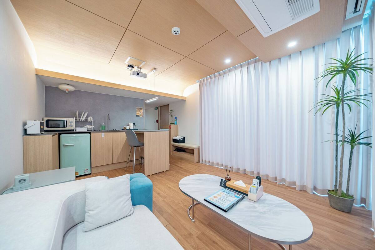 Una habitación de hotel moderna con kitchenette, una zona de estar con sofá y una mesita, y un ventanal con cortinas.