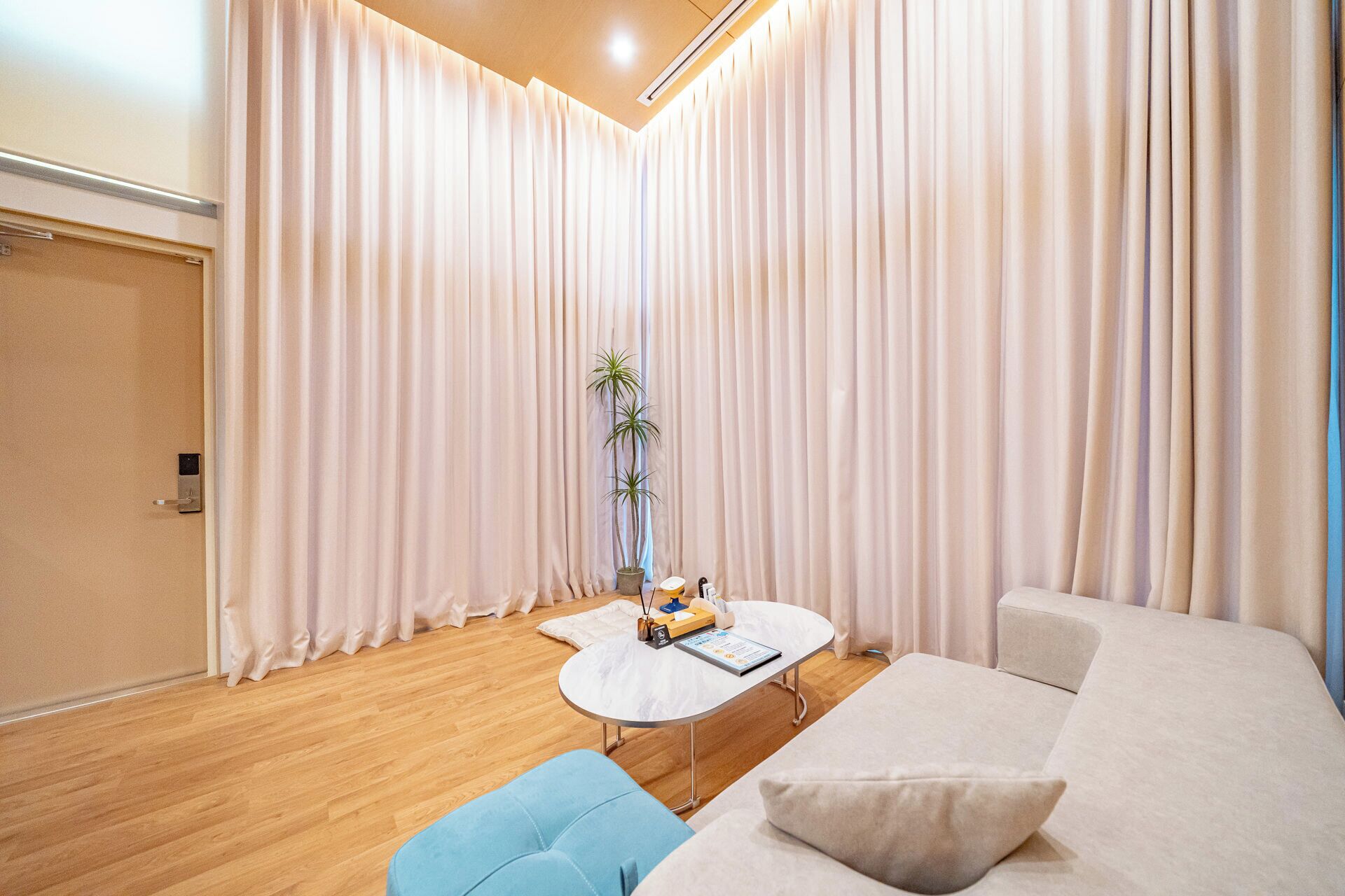 Une chambre d’hôtel moderne avec un plancher en bois, un canapé blanc, une table basse ronde et une grande fenêtre ornée de rideaux légers.