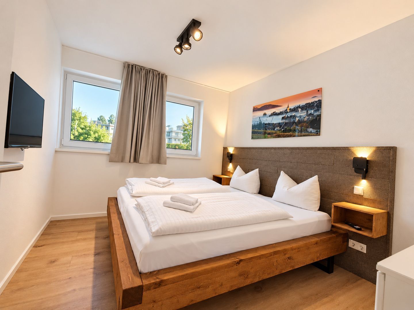 Un lit double avec un cadre en bois, deux tables de chevet avec des lampes, un téléviseur à écran plat fixé au mur, une fenêtre avec des rideaux et un tableau au mur.