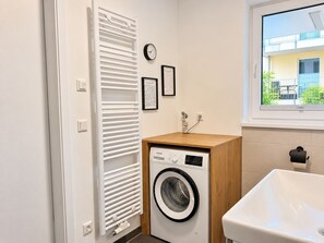 A compact laundry room with a front-loading washing machine, a white radiator, a small sink, and a window with a view of a building outside.
