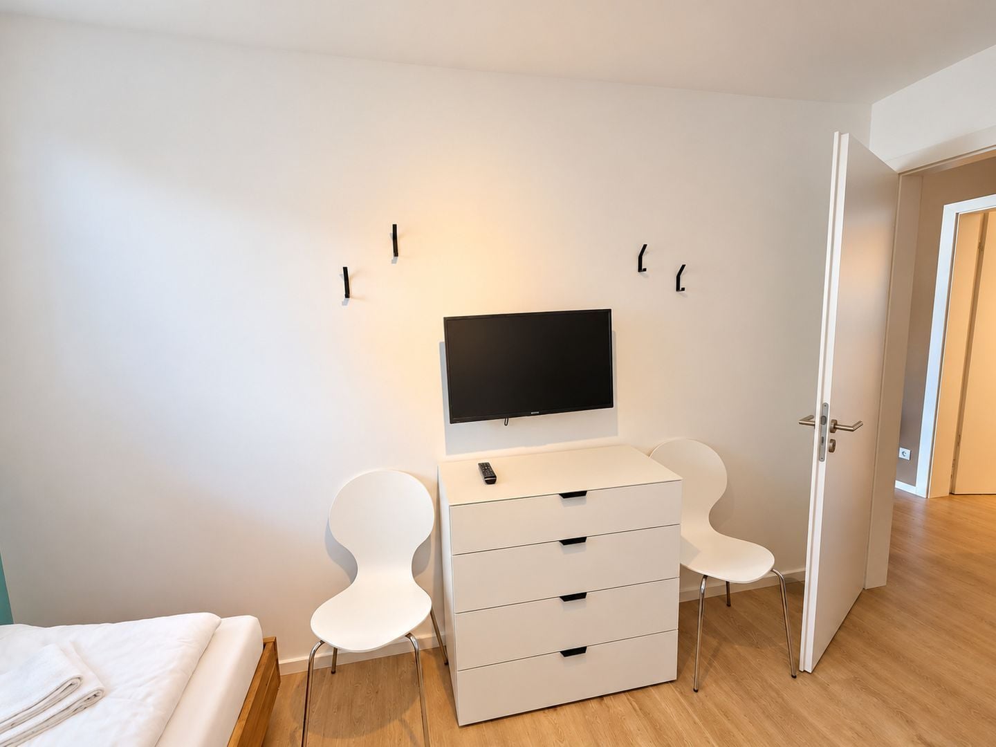 Une chambre moderne avec un lit, un bureau doté d’un téléviseur à écran plat, deux chaises blanches et une commode blanche.