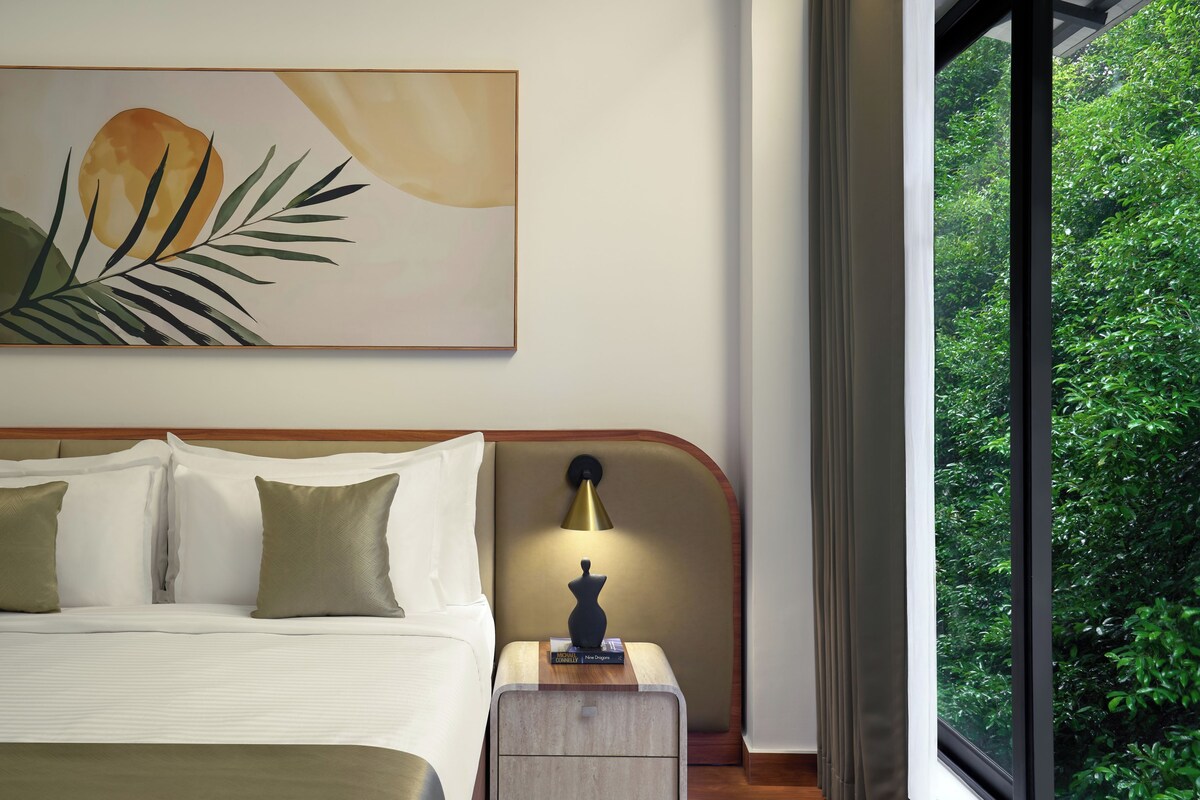 Una camera d'albergo con un letto, cuscini, un comodino con una lampada, un'opera d'arte incorniciata e una finestra vista verde.