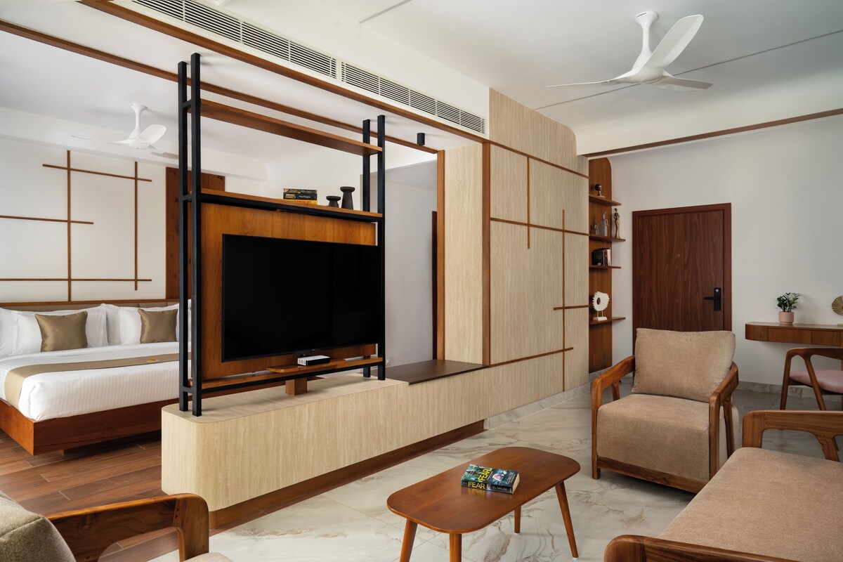 A modern hotel room with a large flat-screen TV, a bed, a sofa, a wooden coffee table, and a desk with a chair.