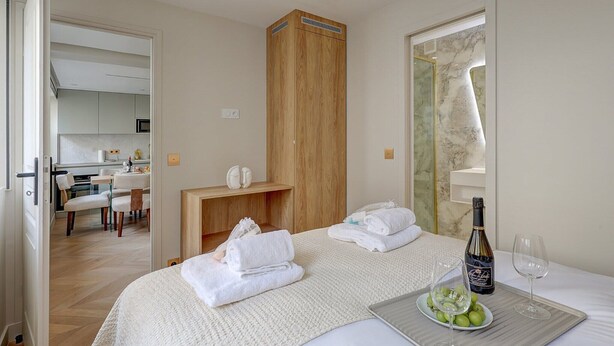 A hotel room with a bed, a wooden wardrobe, a small table with towels, a bottle of wine, and a plate of grapes. - Sublime AC Apartment Eiffel Tower Champs de Mars (Paris)