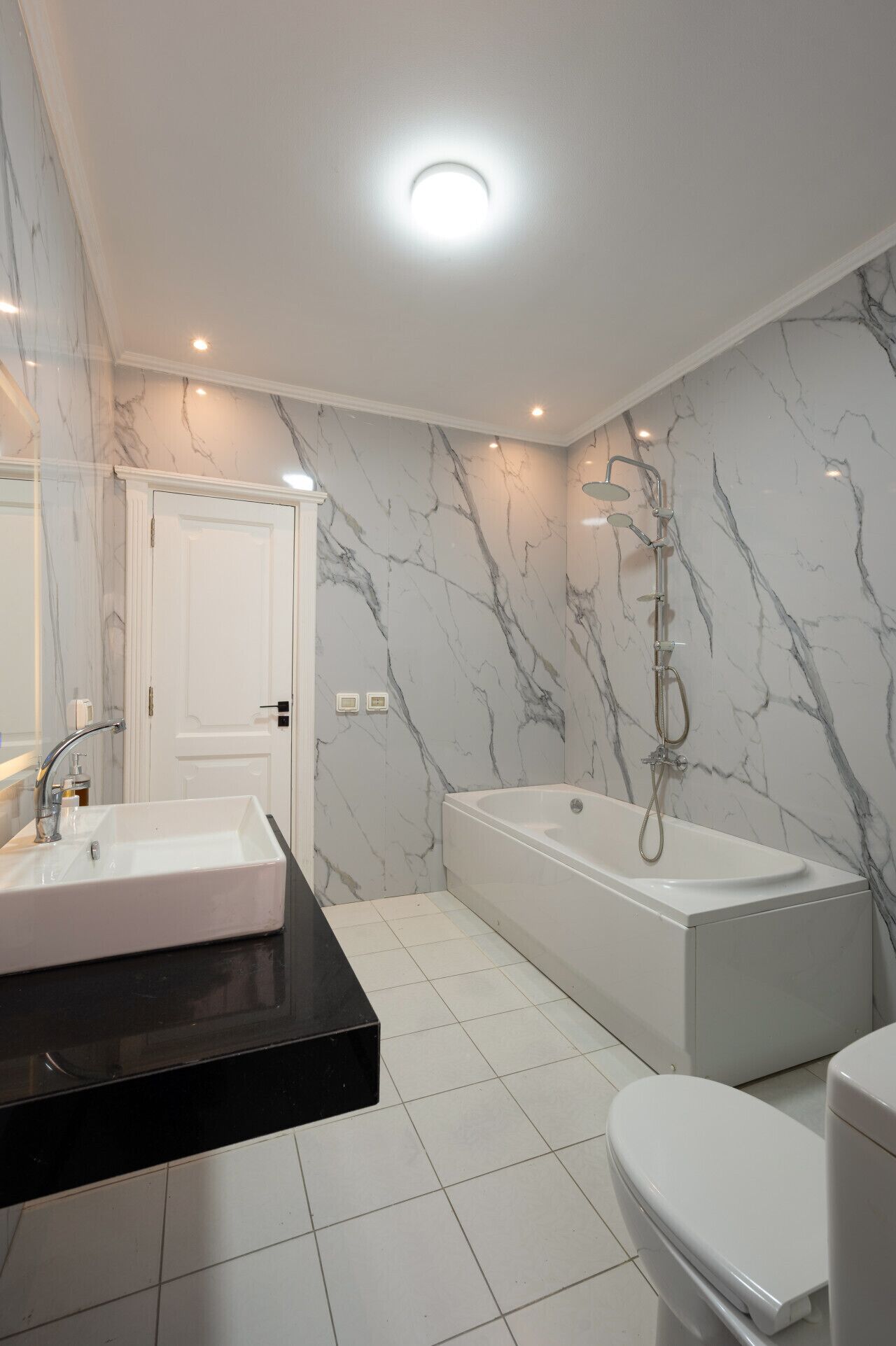 Une salle de bain moderne avec un mur en marbre blanc, un plan de toilette noir, un lavabo blanc, une baignoire et des toilettes.