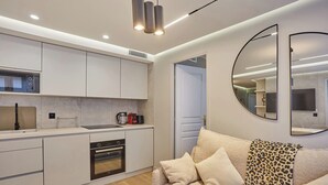 Une cuisine moderne avec des meubles blancs, un four encastré et une machine à café. On y trouve un canapé recouvert d’un plaid à motifs léopard et un miroir rond accroché au mur.