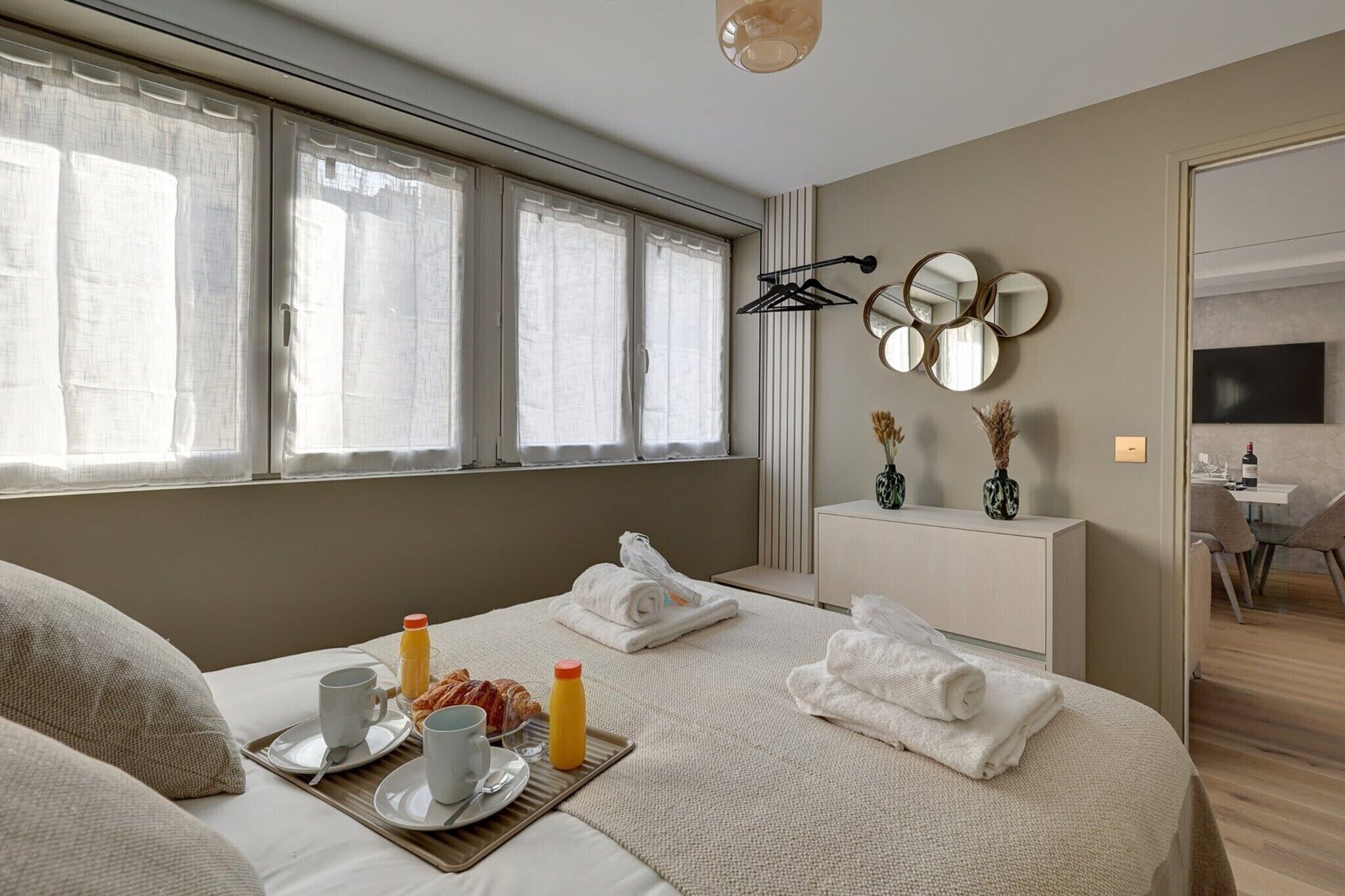 A hotel room with a bed, breakfast tray, towels, a bedside table with decorative items, a TV, and a window with curtains.