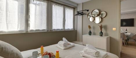 A hotel room with a bed, breakfast tray, towels, a bedside table with decorative items, a TV, and a window with curtains.