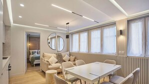 Une pièce moderne et bien éclairée, avec une grande table et des chaises, un canapé recouvert d'un plaid à motifs léopard, un grand miroir rond et de larges fenêtres avec des rideaux.