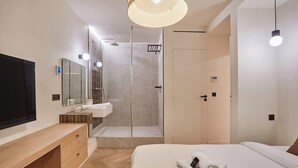 Ein modernes Hotelzimmer mit einem großen Bett, einem Nachttisch aus Holz, einem integrierten Fernseher, einer Duschkabine und einem großen Spiegel.