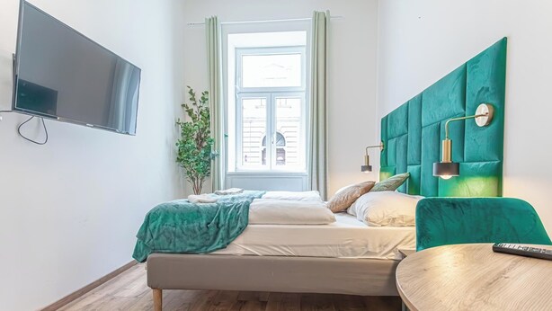 A modern bedroom with a bed, a TV mounted on the wall, a window with curtains, and a green upholstered headboard. - Cozy 1BR Apartment in Prime Location (Vienna)