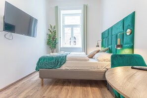 Un dormitorio moderno con cama, televisor montado en la pared, ventana con cortinas y cabecera tapizada de color verde.